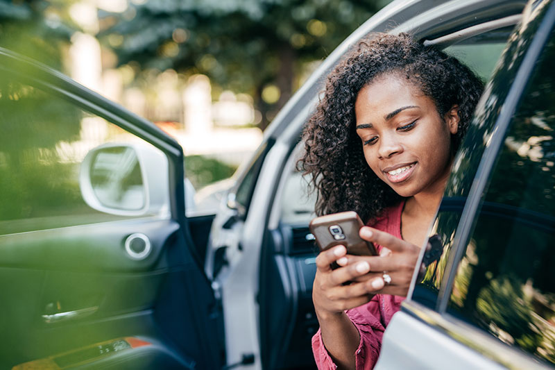 woman sitting in her parked car with the door open using her phone